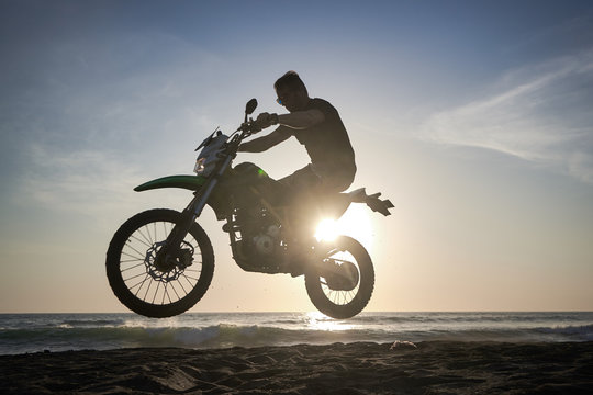 Man on the motorbike mke jump on the black sand beach against sun