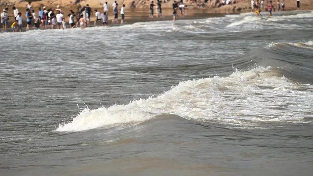 Close up of wave on muddy beach with crowded tourists background, camera pan, high speed, super slow motion, 180 FPS.