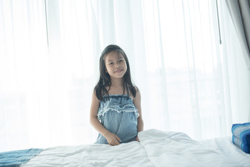 A bright, cute girl stood beside the bed in her bedroom. Beautiful little girl smiling. Portrait of cheerful kid lies at windowsill.