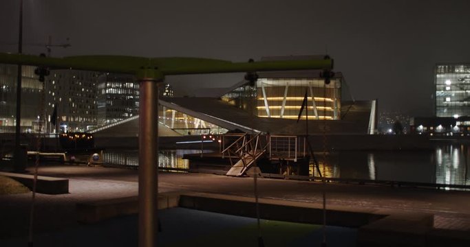 Cinematic wide angle 4K shot with slow parallax motion of empty swings on playground in front of golden illuminated Opera and Music House and Edward Munch Gallery Lambda, at night in Oslo Norway.