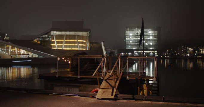 Cinematic wide angle 4K shot with slow parallax motion of sauna in front of golden illuminated Opera House and Edvard Munch gallery Lambda, with light reflection in the fjord, at night in Oslo Norway.