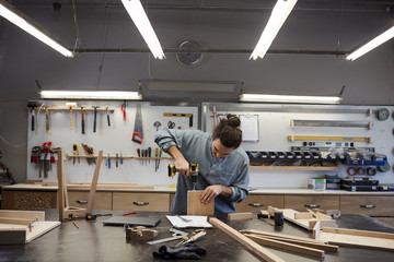 Young carpenter standing at the table and working with wooden detail and drill in workshop