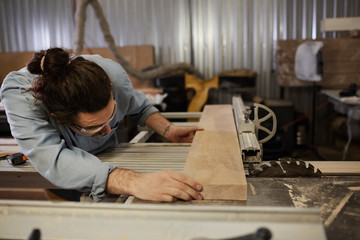Young manual worker in protective eyeglasses fitting the wooden plank and cutting it on lathe in...