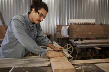 Serious carpenter in protective eyeglasses grinding the wooden plank on lathe in workshop