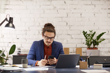 Serious young designer in eyeglasses sitting at the table in front of laptop and working online on...