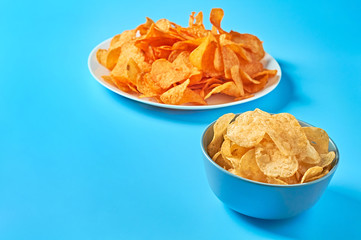 White ceramic plate and bowl full of potato chips on blue background. Space for text