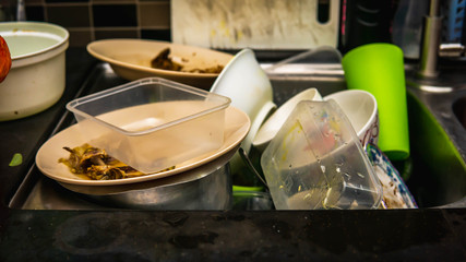 Dirty dishes, utensil, plastic container and food waste piled in a metal sink at the kitchen.