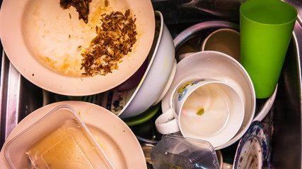 Dirty dishes, utensil, plastic container and food waste piled in a metal sink at the kitchen.