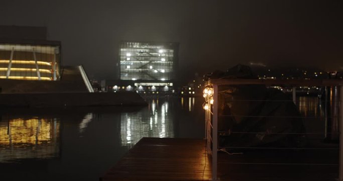 Wide angle 4K shot with slow parallax reveal motion of a sauna with golden illuminated Opera Music and Ballet House and Edvard Munch Gallery Museum in background, at night in Bj&oslash;rvika Oslo Norway.