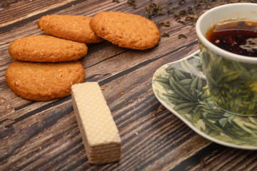 A Cup of black tea, tea leaves, pieces of brown sugar, oatmeal cookies, waffles on a wooden background. Close up.