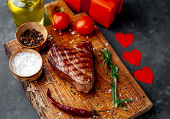 grilled beef steak in the form of a heart on a chopping board with spices and a gift with a red ribbon and hearts for Valentine's day on a stone background with copy space for your text