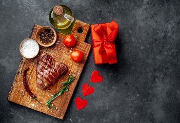 Fototapeta na wymiar grilled beef steak in the form of a heart on a chopping board with spices and a gift with a red ribbon and hearts for Valentine's day on a stone background with copy space for your text