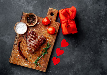 grilled beef steak in the form of a heart on a chopping board with spices and a gift with a red ribbon and hearts for Valentine's day on a stone background with copy space for your text