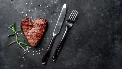 Poster grilled beef steak in the form of a heart with spices, a knife and a fork for Valentine's Day on a stone background with copy space for your text © александр таланцев