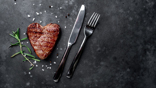 grilled beef steak in the form of a heart with spices, a knife and a fork for Valentine's Day on a stone background with copy space for your text