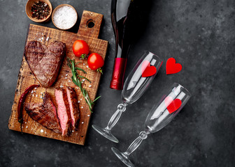 Fototapeta na wymiar two grilled beef steaks in the form of a heart with spices, a bottle of champagne or wine with two glasses for dinner for Valentine's day on a stone background with copy space for your text