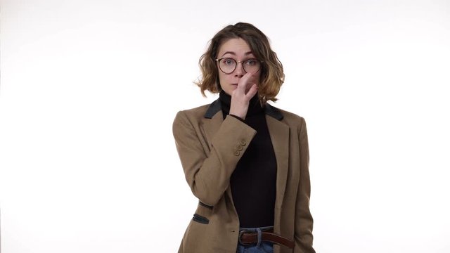 Curly haired european woman in stylish eyeglasses looking to the camera and showing silence gesture, keeping secrets. Metaphorically fastens her mouth and throws a key. Cheerfully smiling