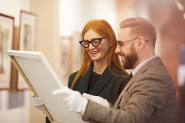 Young woman in eyeglasses holding picture and looking at it together with young man they standing and smiling in gallery