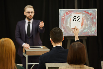 Rear view of young man raising his sign with number he is going to buy a painting at auction