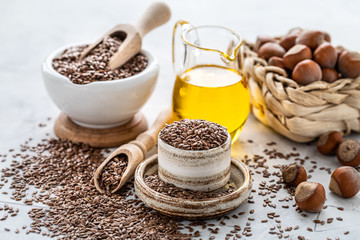 Walnut and flaxseed oil in a bottle and ceramic bowl with brown flax seeds and a wooden spoon on a white background.