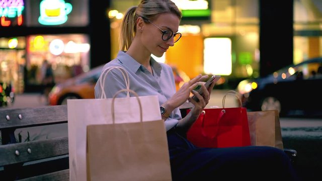 Happy female typing funny content publication with life hacks for shopping with discounts using public internet connection in downtown, positive woman with paper packages resting with cellular gadget