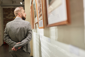 Rear view of young man in suit looking at paintings on the wall while walking along the corridor of...