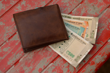 Colorful new Indian Rupee notes of fifty, two and five hundred peeking out of a fat brown color leather wallet kept outdoor on a red park bench. Concept shot depicting inflation and rich people.