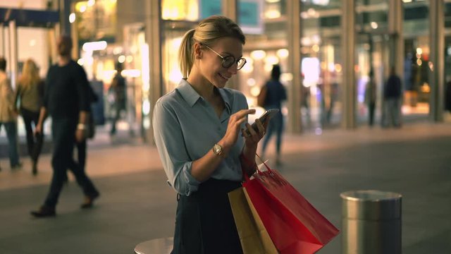Happy woman with purchase bags searching information about shopping sales received funny text message from friend, cheerful hipster girl reading positive news on fashion website and laughing