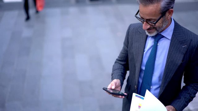 Successful man in optical spectacles for vision correction typing text message via cellphone application while doing accounting with financial report, concept of browsing network system