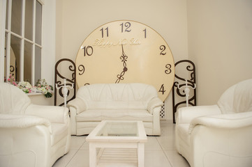 close up photo of a big round retro clock in a living room with beige leather sofas and a table between them