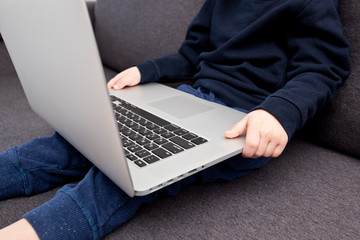 The child is sitting on the sofa holding the laptop and watches the cartoons