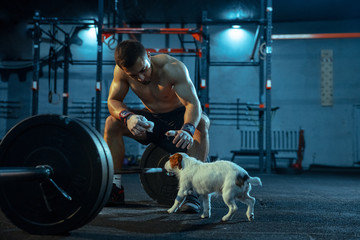 Caucasian man practicing in weightlifting in gym. Male sportive model resting after training, looks strong, petting dog. Body building, healthy lifestyle, movement, activity, action concept.