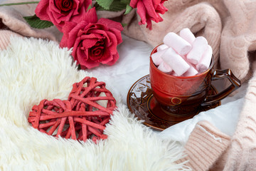 Bouquet red rose flowers and wicker red heart on white blanket, cup with marshmallow and pink sweater. Beautiful background for Valentine's Day or wedding. Lifestyle. Top view, flat lay.