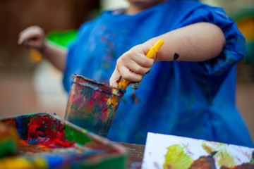 Child at nursery painting with a brush wearing a painting apron with paint all over it. Close up of...