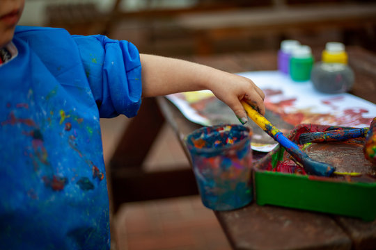 Child At Nursery Painting With A Brush And Roller, Learning Colour And Creativity Through Messy Play. Close Up Of The Paints And The Child Holding A Paint Brush To Create A Painting.