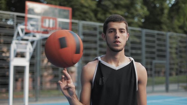 Close Up of Young Caucasian Basketball Player Wearing Black Singlet Spinning the Ball on his Finger Standing at Street Basketbal Court and Looking to Camera. Healthy Lifestyle and Sport Concept.