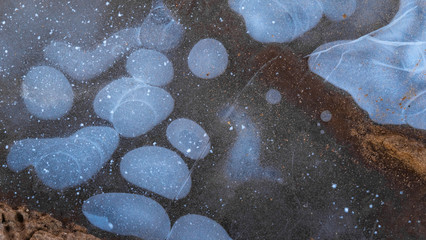Frozen puddle. The surface of frozen water in a small puddle, snow and sand lie on top.Closeup.Air frozen in ice creates white bubbles.Countryside. The first autumn frosts.Temperature change. 