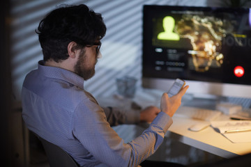 Rear view of bearded manager using his mobile phone while sitting at the table with computer and has online conference at office