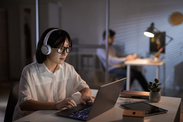 Serious Asian designer in headphones working on laptop computer at the office till late evening