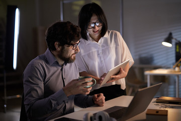 Asian businesswoman showing online presentation on tablet pc to her colleague and they discussing it together at dark office