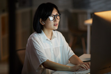 Serious Asian businesswoman wearing eyeglasses looking at computer monitor and typing she working...