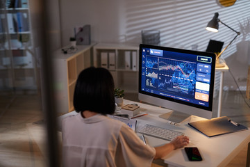 Rear view of young analyst sitting at the table in front of computer monitor with financial charts...
