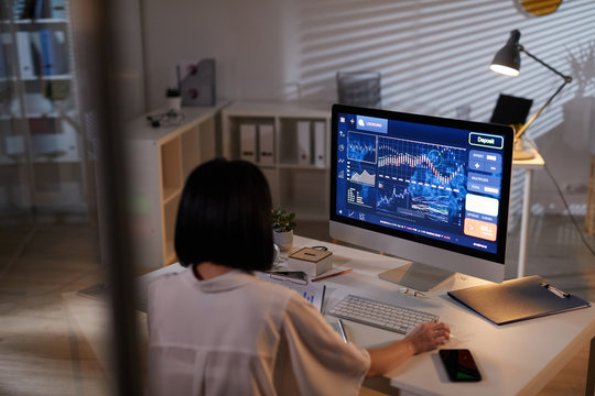 Rear View Of Young Analyst Sitting At The Table In Front Of Computer Monitor With Financial Charts And Analyzing It At Office