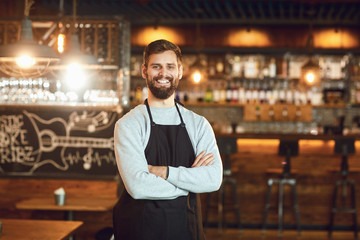 Bearded smiling barman waiter standing on the background of a bar. - obrazy, fototapety, plakaty