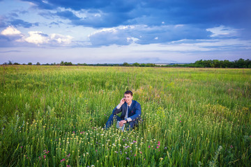 Fototapeta premium Businessman sitting in his suit on a green sunny field phoning from his mobile phone