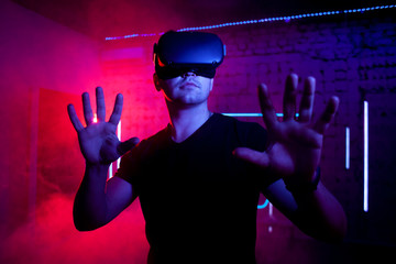 serious brutal guy with VR glasses stands in the virtual reality room and plays with joysticks,...