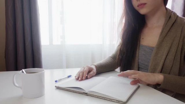 A young woman sits at a table and writes notes in a notebook. A list of important tasks for the week. Close-up