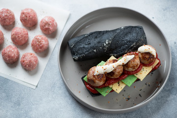 Sub with meatballs and cheese served on a grey plate, elevated view over light-blue stone surface with uncooked meatballs