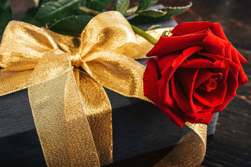 Gift wrapped in black paper and a gold ribbon. on top of the box is a luxurious red rose. The concept of an expensive gift for the holiday Valentine's Day or Women's Day March 8