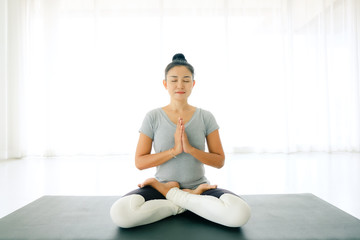 Asian woman sitting on mat and doing yoga and raise hand or pay(namaste) obeisance with  legs crossed and eyes closed in room at home,Concept of healing body and spirit.